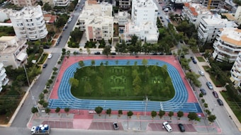 An aerial view of a sports complex featuring a blue running track surrounding a green field with workout equipment and trees. The area is bordered by residential buildings and streets with parked cars and greenery.