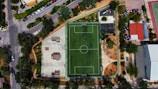 An aerial view of a sports complex featuring a green football field at the center, surrounded by a paved area on the left with skatepark ramps. Surrounding the field are trees, walkways, and small buildings. There's a road with parked cars and nearby green spaces.