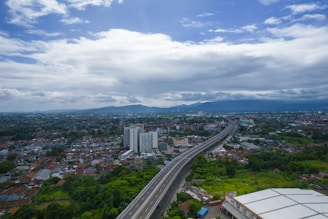 An aerial view of an urban area with a highway cutting through the landscape. The foreground features a large building, possibly an industrial or warehouse property. Residential and commercial buildings are scattered throughout, with greenery interspersed. The highway leads towards a cityscape in the distance, with mountains visible under a mostly cloudy sky.