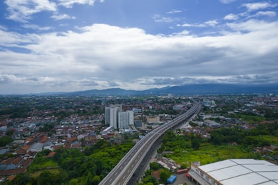 An aerial view of an urban area with a highway cutting through the landscape. The foreground features a large building, possibly an industrial or warehouse property. Residential and commercial buildings are scattered throughout, with greenery interspersed. The highway leads towards a cityscape in the distance, with mountains visible under a mostly cloudy sky.