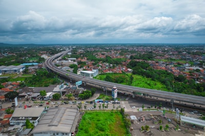 An aerial view of a sprawling urban area with a prominent highway cutting through the landscape. Residential houses with red-tiled roofs densely populate the area on both sides of the highway. Lush green patches suggest parks or undeveloped land. The sky is overcast with thick clouds, adding depth to the horizon.