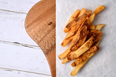 A pile of crispy golden French fries is placed on a piece of parchment paper. The fries have a rustic appearance with some skin left on. To the left, there is a wooden surface with visible grain.