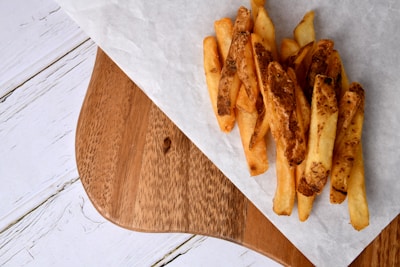 A portion of rustic-style French fries with skins on, placed on a piece of white parchment paper. The fries rest on a wooden serving board, which contrasts with a white, slightly textured background surface.