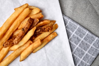 A pile of golden-brown French fries is arranged on a piece of parchment paper atop a textured surface. The surrounding area features gray and white woven cloths, adding a rustic and homey feel.