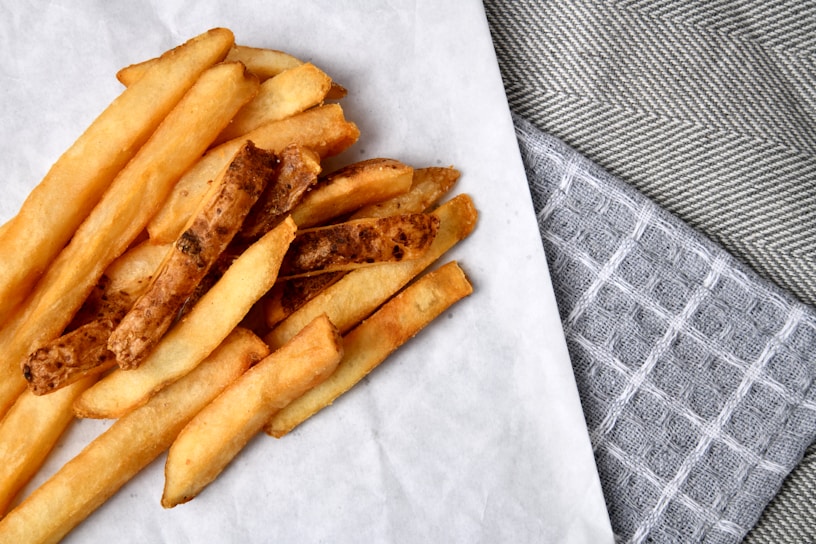A pile of golden-brown French fries is arranged on a piece of parchment paper atop a textured surface. The surrounding area features gray and white woven cloths, adding a rustic and homey feel.