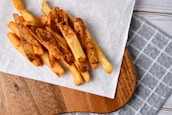 A portion of golden brown French fries rests on a piece of white parchment paper placed on a wooden serving board. The board sits on a grey and white checkered cloth, adding a rustic touch to the presentation. The fries look crispy and freshly prepared.