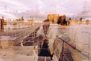 A reflective scene capturing a person holding a camera, with their image superimposed on a waterfront backdrop. The setting includes a boardwalk or pier with railings and a view of buildings in the distance, enhanced by warm evening or sunset lighting.