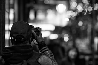 A person with a cap is holding a camera, aiming to capture a scene. The background contains blurred lights, creating a bokeh effect, giving the scene a lively, urban atmosphere.
