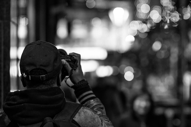 A person with a cap is holding a camera, aiming to capture a scene. The background contains blurred lights, creating a bokeh effect, giving the scene a lively, urban atmosphere.