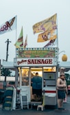 A small food stand is selling items like Italian sausage, fresh cut fries, and cheesesteaks. The stand is adorned with colorful signs and flags. A person is inside the illuminated food truck preparing food. There is a humorous statue of a chef standing beside the stand. Various items like folding chairs and cleaning supplies are placed next to the entrance.