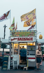 A small food stand is selling items like Italian sausage, fresh cut fries, and cheesesteaks. The stand is adorned with colorful signs and flags. A person is inside the illuminated food truck preparing food. There is a humorous statue of a chef standing beside the stand. Various items like folding chairs and cleaning supplies are placed next to the entrance.