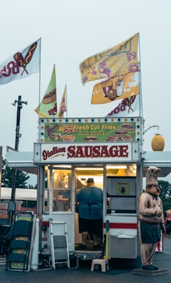 A small food stand is selling items like Italian sausage, fresh cut fries, and cheesesteaks. The stand is adorned with colorful signs and flags. A person is inside the illuminated food truck preparing food. There is a humorous statue of a chef standing beside the stand. Various items like folding chairs and cleaning supplies are placed next to the entrance.