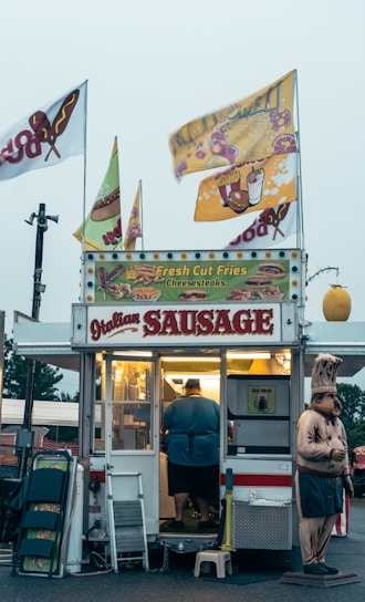A small food stand is selling items like Italian sausage, fresh cut fries, and cheesesteaks. The stand is adorned with colorful signs and flags. A person is inside the illuminated food truck preparing food. There is a humorous statue of a chef standing beside the stand. Various items like folding chairs and cleaning supplies are placed next to the entrance.