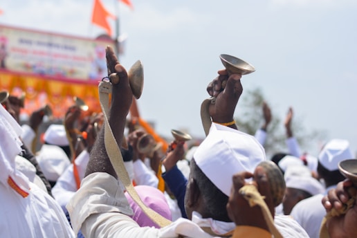 A group of people dressed in white clothing, holding small metallic bowls or bells. The background features vibrant orange and red colors, possibly from banners or flags. The scene appears festive and communal, with hands raised skyward, engaging in some form of collective activity.