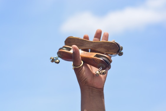 A hand is holding a traditional wooden musical instrument with several small bells attached. The object is being held up against a clear blue sky.
