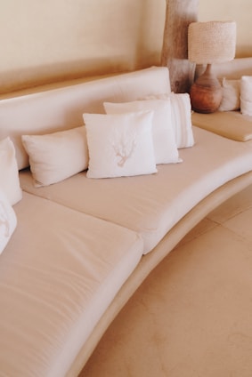 A minimalist, neutral-toned living space featuring a curved sofa adorned with several white decorative pillows. A rustic wooden lamp with a textured shade sits at one end of the sofa on a smooth, light-colored floor.