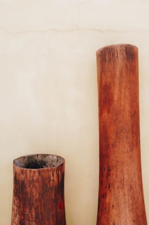 Two large, rustic wooden vases with a weathered texture stand against a plain beige wall. The vases have a cylindrical shape with a natural wood grain that fades from a dark brown at the base to a lighter brown at the top.