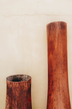 Two large, rustic wooden vases with a weathered texture stand against a plain beige wall. The vases have a cylindrical shape with a natural wood grain that fades from a dark brown at the base to a lighter brown at the top.