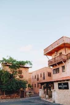 A rustic building with clay-colored walls and wooden balconies is set against a clear blue sky. The structure is complemented by lush greenery and cobblestone streets, giving a quaint and historic vibe. A sign reading 'El Mercadito' is displayed, adding a sense of place.