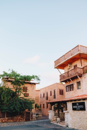 A rustic building with clay-colored walls and wooden balconies is set against a clear blue sky. The structure is complemented by lush greenery and cobblestone streets, giving a quaint and historic vibe. A sign reading 'El Mercadito' is displayed, adding a sense of place.