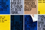 A grid of posters featuring bold, geometric typography in various colors. Some posters have words that are partially obscured or peeled. The background colors include blue, yellow, and beige, with different text orientations. One poster appears to be partially peeled back to reveal a layer underneath.