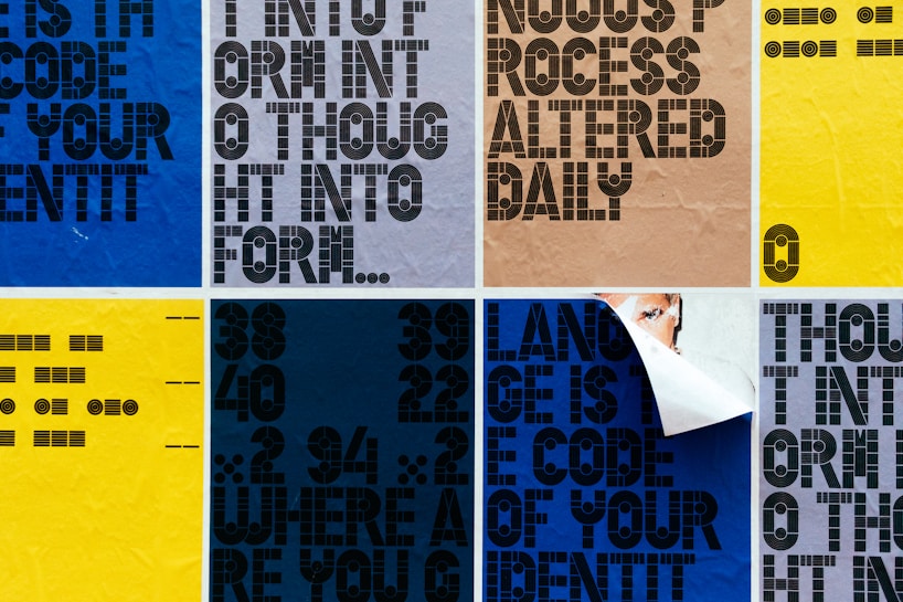 A grid of posters featuring bold, geometric typography in various colors. Some posters have words that are partially obscured or peeled. The background colors include blue, yellow, and beige, with different text orientations. One poster appears to be partially peeled back to reveal a layer underneath.