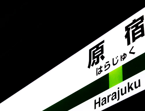 A sign containing both Japanese characters and Roman letters spelling 'Harajuku', set against a predominantly dark background with a green stripe.