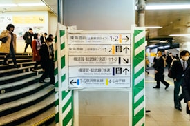 A busy underground station with various people walking in different directions. Multiple directional signs in Japanese and English guide commuters to different train lines, including Tokaido Line, Yokosuka Line, and Yamanote Line. The atmosphere is structured, with clear pathways and staircases leading up and down.