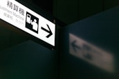 A sign with an illustration of a person interacting with a machine and the text 'Adjustment Machine'. The sign is illuminated and casts a shadow on a nearby wall. The setting appears to be in a public place, possibly a transportation hub.