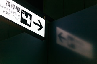 A sign with an illustration of a person interacting with a machine and the text 'Adjustment Machine'. The sign is illuminated and casts a shadow on a nearby wall. The setting appears to be in a public place, possibly a transportation hub.