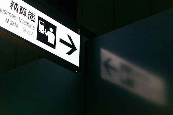 A sign with an illustration of a person interacting with a machine and the text 'Adjustment Machine'. The sign is illuminated and casts a shadow on a nearby wall. The setting appears to be in a public place, possibly a transportation hub.