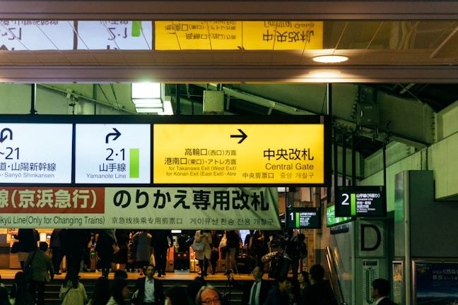 A busy train station with multiple signs in different languages directing to platforms and gates. The atmosphere is bustling with passengers navigating through the space. The signage includes directions for the Yamanote Line and the Central Gate.