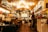 A cozy restaurant or café with warm lighting and a welcoming atmosphere. Several people are seated at tables, enjoying their meals and conversation. The interior features wooden décor, colorful lights, and vibrant signs. A counter area is visible with staff preparing orders.