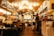 A cozy restaurant or café with warm lighting and a welcoming atmosphere. Several people are seated at tables, enjoying their meals and conversation. The interior features wooden décor, colorful lights, and vibrant signs. A counter area is visible with staff preparing orders.
