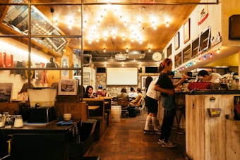 A cozy restaurant or café with warm lighting and a welcoming atmosphere. Several people are seated at tables, enjoying their meals and conversation. The interior features wooden décor, colorful lights, and vibrant signs. A counter area is visible with staff preparing orders.