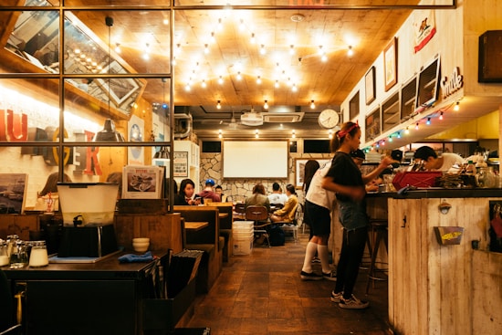 A cozy restaurant or caf&eacute; with warm lighting and a welcoming atmosphere. Several people are seated at tables, enjoying their meals and conversation. The interior features wooden d&eacute;cor, colorful lights, and vibrant signs. A counter area is visible with staff preparing orders.