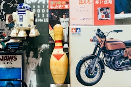A collection of vintage items is arranged closely together, including an R2-D2 model, a large bowling pin, a Tamiya model motorcycle box, and several vintage posters, such as one featuring the film Jaws and a photograph of a person in a Giants baseball uniform. The scene mixes elements of pop culture and nostalgia.