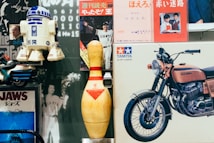 A collection of vintage items is arranged closely together, including an R2-D2 model, a large bowling pin, a Tamiya model motorcycle box, and several vintage posters, such as one featuring the film Jaws and a photograph of a person in a Giants baseball uniform. The scene mixes elements of pop culture and nostalgia.