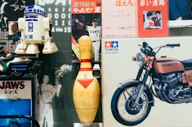 A collection of vintage items is arranged closely together, including an R2-D2 model, a large bowling pin, a Tamiya model motorcycle box, and several vintage posters, such as one featuring the film Jaws and a photograph of a person in a Giants baseball uniform. The scene mixes elements of pop culture and nostalgia.