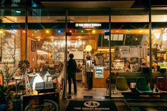 A cozy, warmly lit burger restaurant with a retro American diner theme. The interior is lively with people dining at tables and the counter. The decor includes colorful lights, American flags, and various signs. The front window displays the name 'The Great Burger Tokyo Japan'.