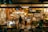 A cozy, warmly lit burger restaurant with a retro American diner theme. The interior is lively with people dining at tables and the counter. The decor includes colorful lights, American flags, and various signs. The front window displays the name 'The Great Burger Tokyo Japan'.