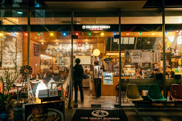 A cozy, warmly lit burger restaurant with a retro American diner theme. The interior is lively with people dining at tables and the counter. The decor includes colorful lights, American flags, and various signs. The front window displays the name 'The Great Burger Tokyo Japan'.