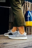 A person is wearing fashionable sneakers with a textured pattern and white soles. The person is standing on a wooden floor, next to a pair of colorful buoys and a wooden fence. Olive green pants partially cover the legs.