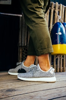 A person is wearing fashionable sneakers with a textured pattern and white soles. The person is standing on a wooden floor, next to a pair of colorful buoys and a wooden fence. Olive green pants partially cover the legs.