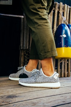 A person is wearing fashionable sneakers with a textured pattern and white soles. The person is standing on a wooden floor, next to a pair of colorful buoys and a wooden fence. Olive green pants partially cover the legs.