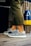A person is wearing fashionable sneakers with a textured pattern and white soles. The person is standing on a wooden floor, next to a pair of colorful buoys and a wooden fence. Olive green pants partially cover the legs.