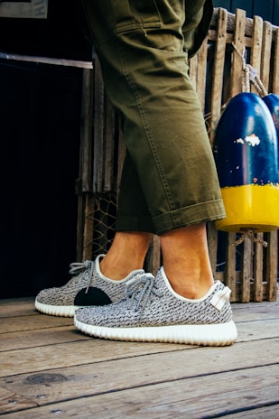 A person is wearing fashionable sneakers with a textured pattern and white soles. The person is standing on a wooden floor, next to a pair of colorful buoys and a wooden fence. Olive green pants partially cover the legs.