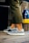A person is wearing fashionable sneakers with a textured pattern and white soles. The person is standing on a wooden floor, next to a pair of colorful buoys and a wooden fence. Olive green pants partially cover the legs.
