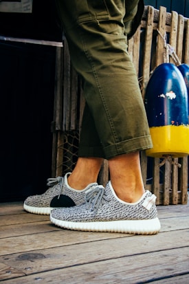 A person is wearing fashionable sneakers with a textured pattern and white soles. The person is standing on a wooden floor, next to a pair of colorful buoys and a wooden fence. Olive green pants partially cover the legs.