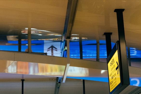 A modern airport terminal with reflective metallic surfaces and bold architectural lines. The yellow directional sign points towards central gates, conveying a sense of movement and direction. Bright blue and chrome reflections add a futuristic feel.
